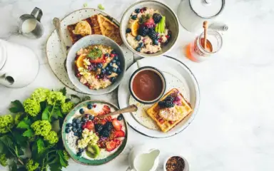 A bright, inviting breakfast spread on a marble table, featuring colorful bowls of oatmeal topped with fruits and seeds, waffles, tea, and a honey jar.