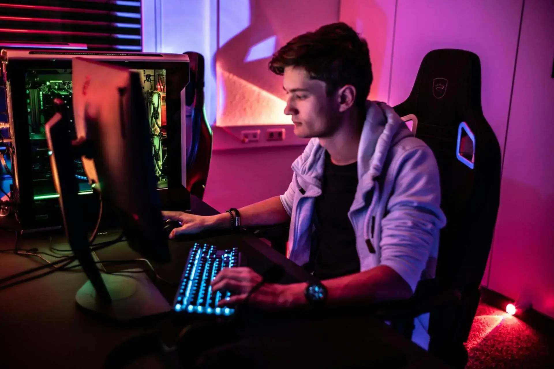 A young man in a hoodie intently uses a computer in a dimly lit room with vibrant neon lights. The scene exudes a focused and modern gaming atmosphere.