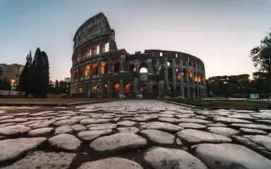 Low-angle view of the Colosseum in Rome at sunset, with its arches illuminated in warm lights. Cobblestone foreground adds historical depth. Serene atmosphere.