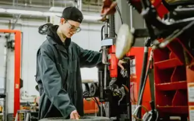 Young male mechanic wearing a cap and safety glasses operates machinery next to a tire in a garage. The setting is industrious and focused.