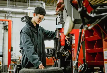 Young male mechanic wearing a cap and safety glasses operates machinery next to a tire in a garage. The setting is industrious and focused.