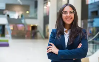 A confident woman in a blue suit stands in a bright, modern atrium, smiling with arms crossed. Background features blurred stairs and glass railings.