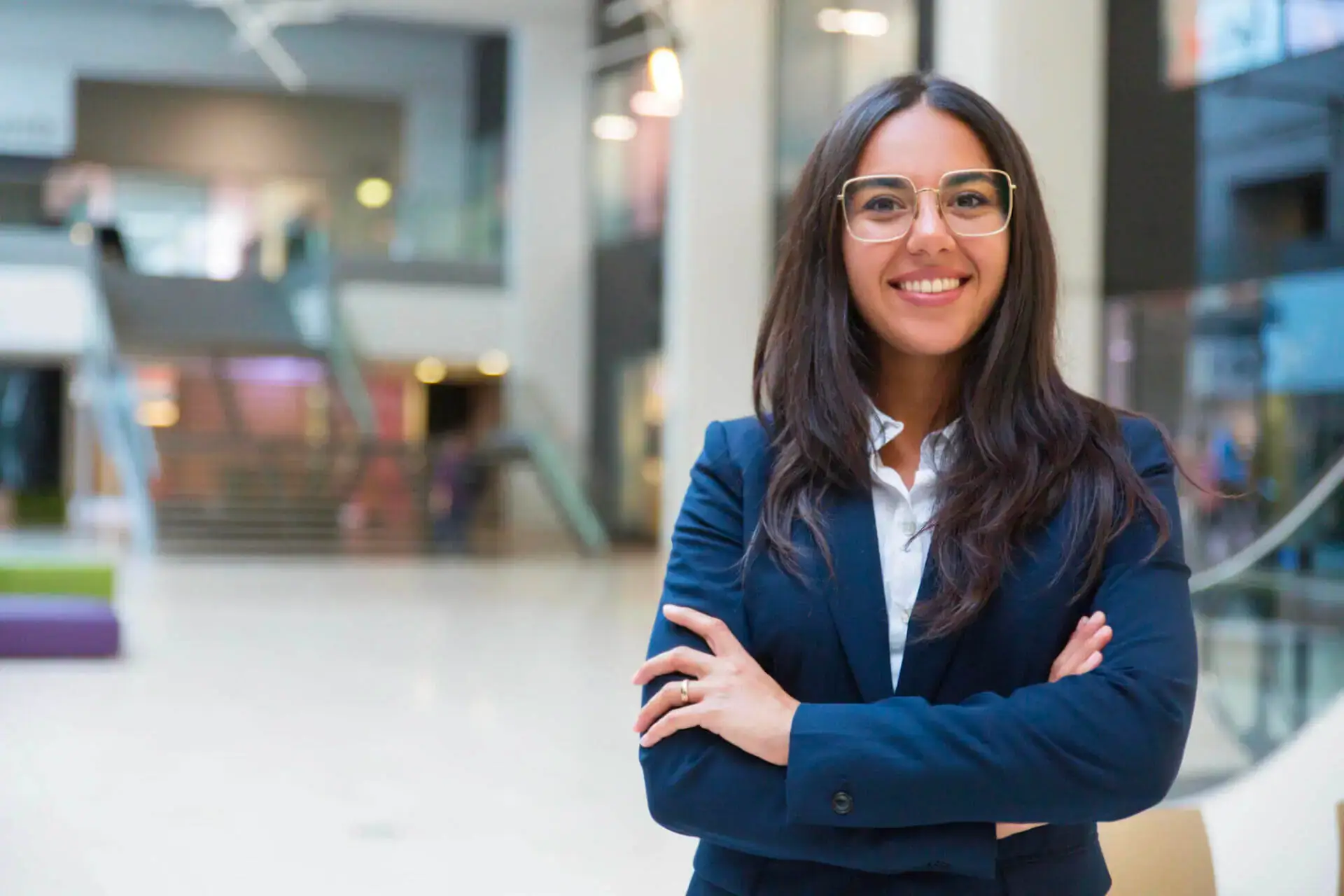 A confident woman in a blue suit stands in a bright, modern atrium, smiling with arms crossed. Background features blurred stairs and glass railings.