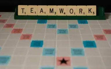 Scrabble tiles spell "TEAMWORK" on a rack above a game board. The board features bonus squares in blue and pink, conveying a collaborative theme.
