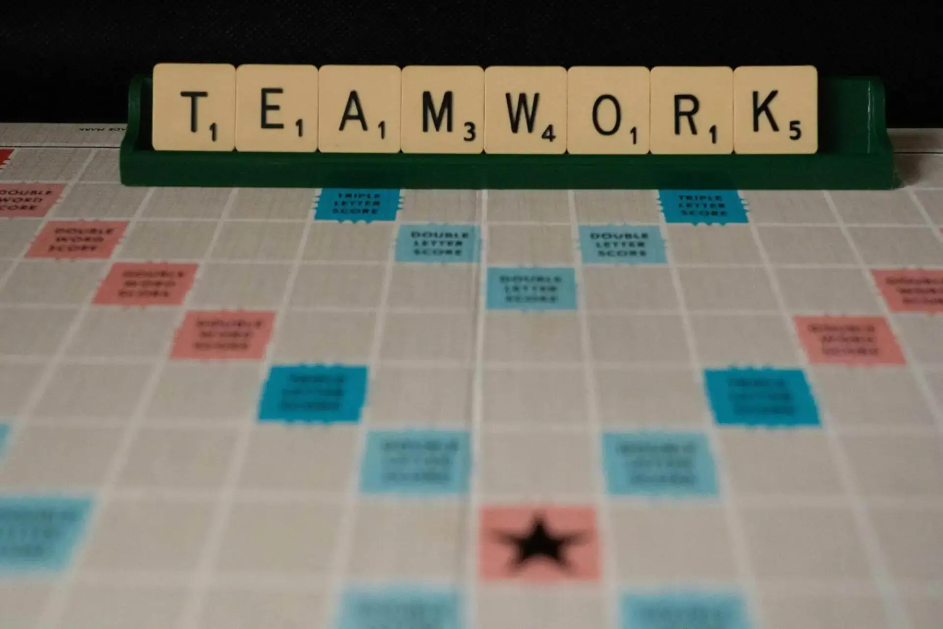 Scrabble tiles spell "TEAMWORK" on a rack above a game board. The board features bonus squares in blue and pink, conveying a collaborative theme.