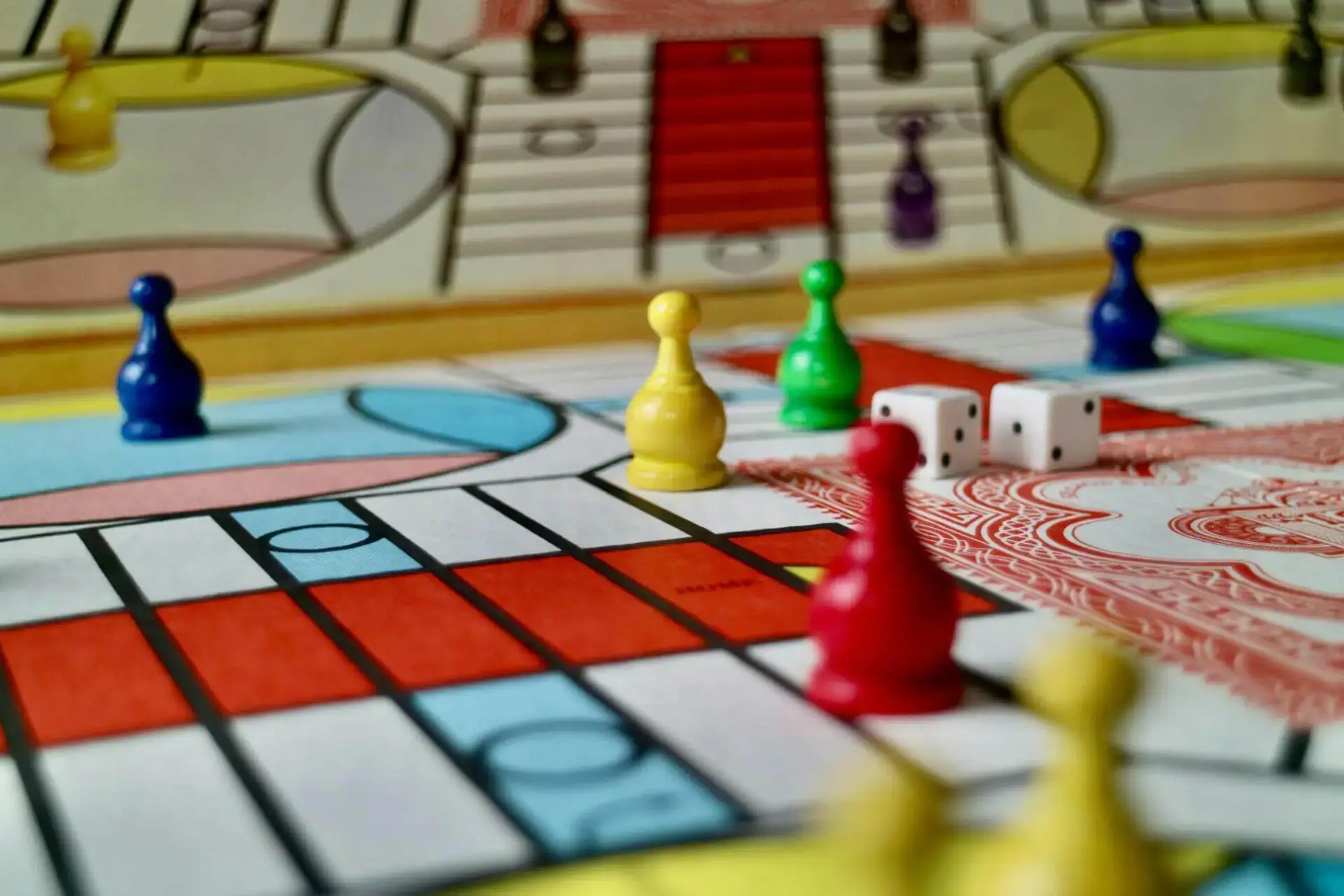 Colorful game board with yellow, red, blue, and green pawns arranged near two white dice. The background shows intricate patterns, suggesting a lively, strategic game.