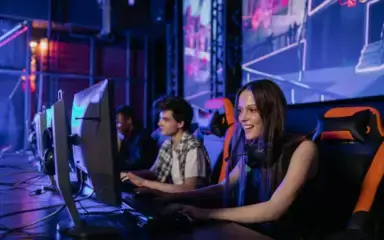 Two people are gaming on computers in a dimly lit room, focused and smiling. The atmosphere is vibrant, with colorful gaming screens displayed.