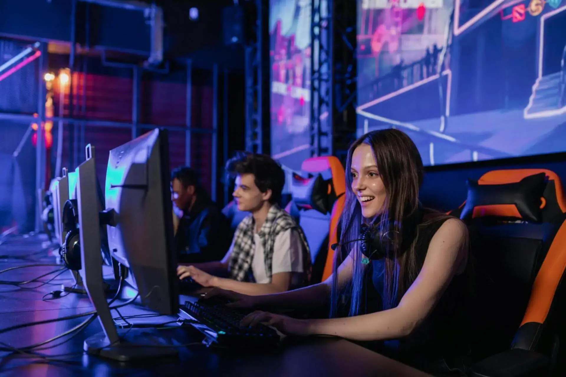 Two people are gaming on computers in a dimly lit room, focused and smiling. The atmosphere is vibrant, with colorful gaming screens displayed.