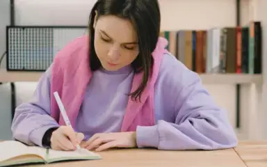 A woman in a lavender sweatshirt and pink scarf writes in a notebook at a wooden table. A bookshelf with books is blurred in the background.