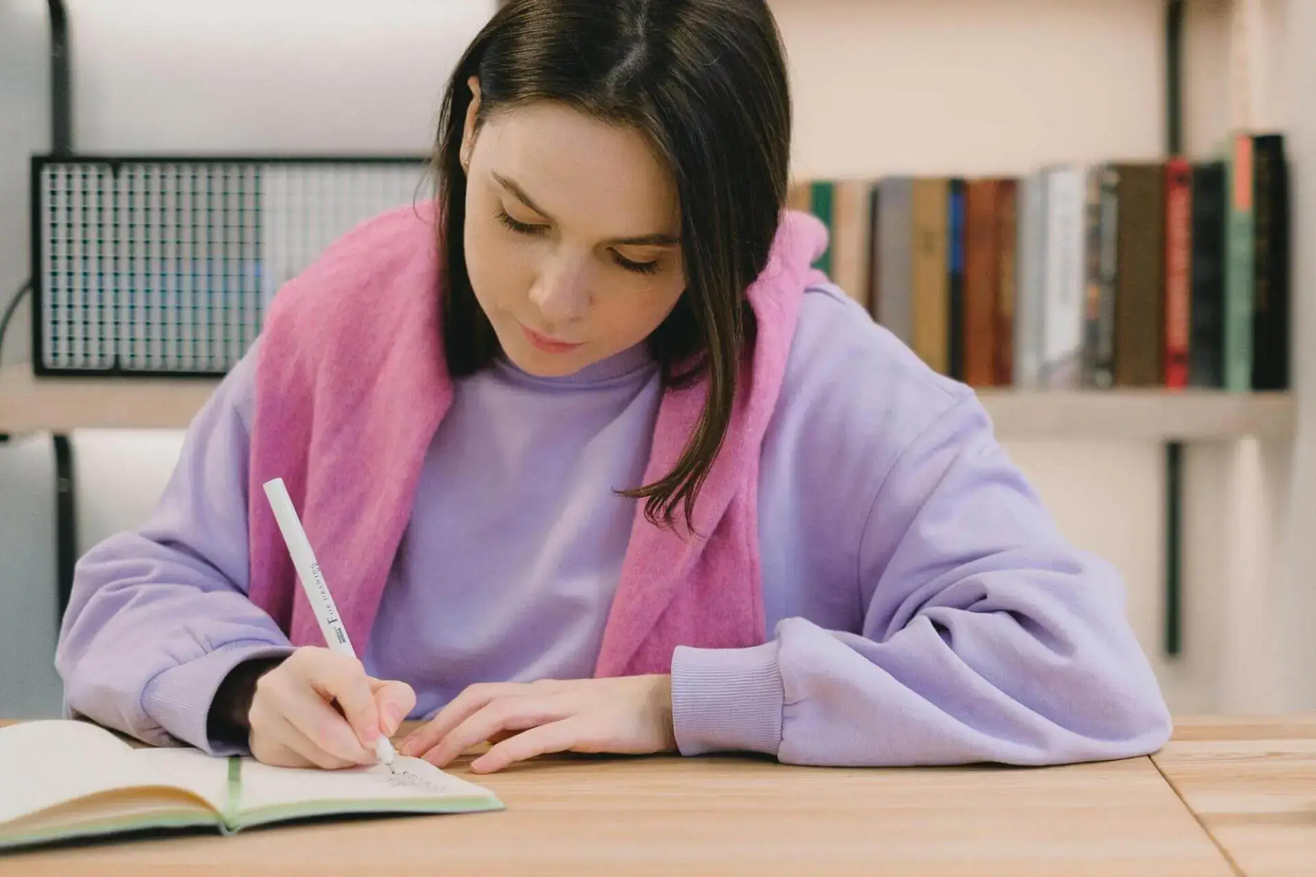 A woman in a lavender sweatshirt and pink scarf writes in a notebook at a wooden table. A bookshelf with books is blurred in the background.