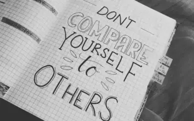 A handwritten motivational quote in a notebook reads, "Don't compare yourself to others," on graph paper. Tabs are visible, adding an organized tone.