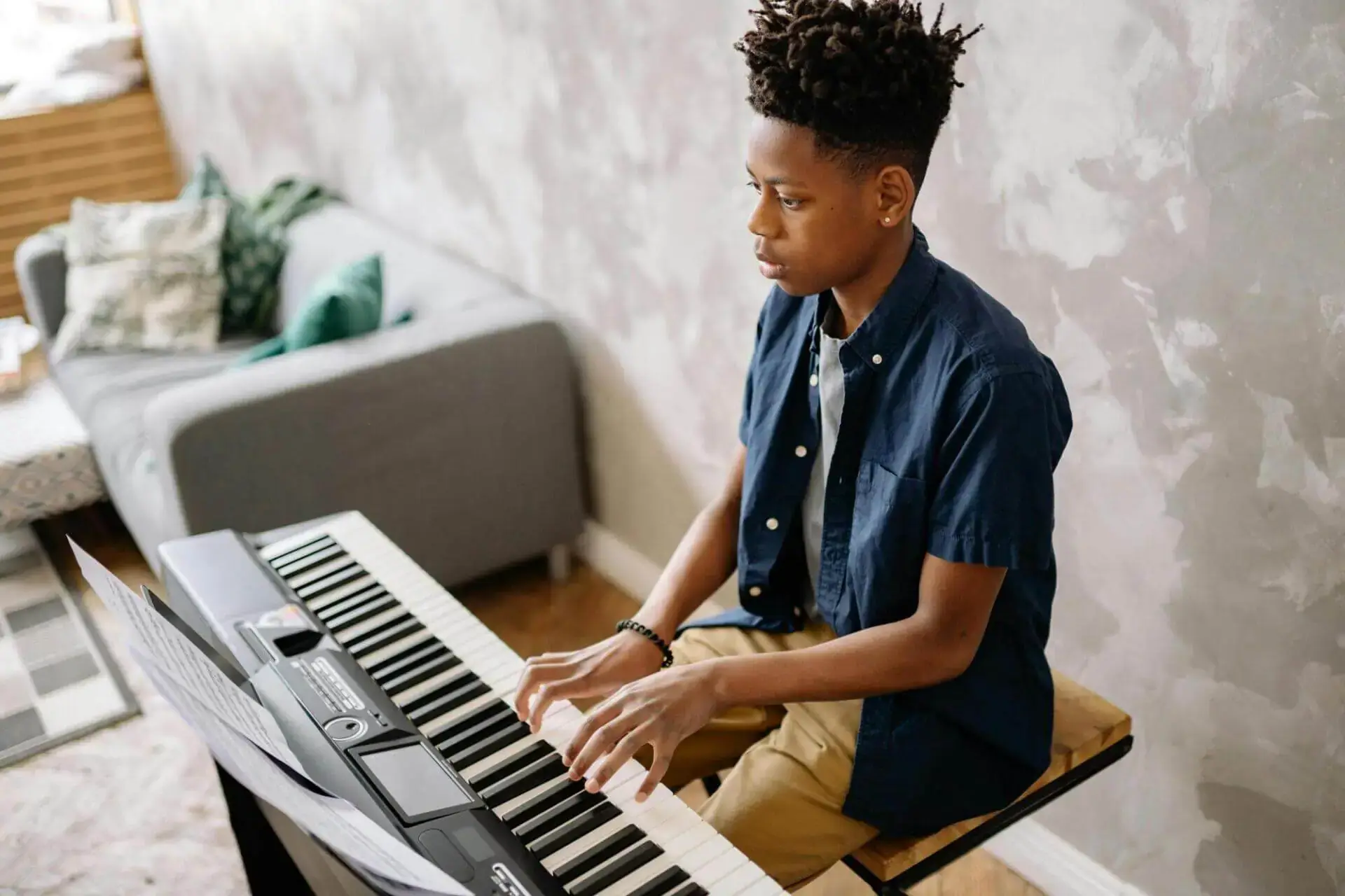 Teen playing a keyboard in a cozy room with a patterned wall and a sofa. The focused expression suggests a serious yet calm atmosphere.