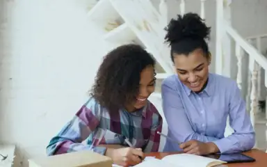 Two women sit at a table, smiling as they engage with an open notebook. The cozy, white background and natural light add a warm, cheerful ambiance.