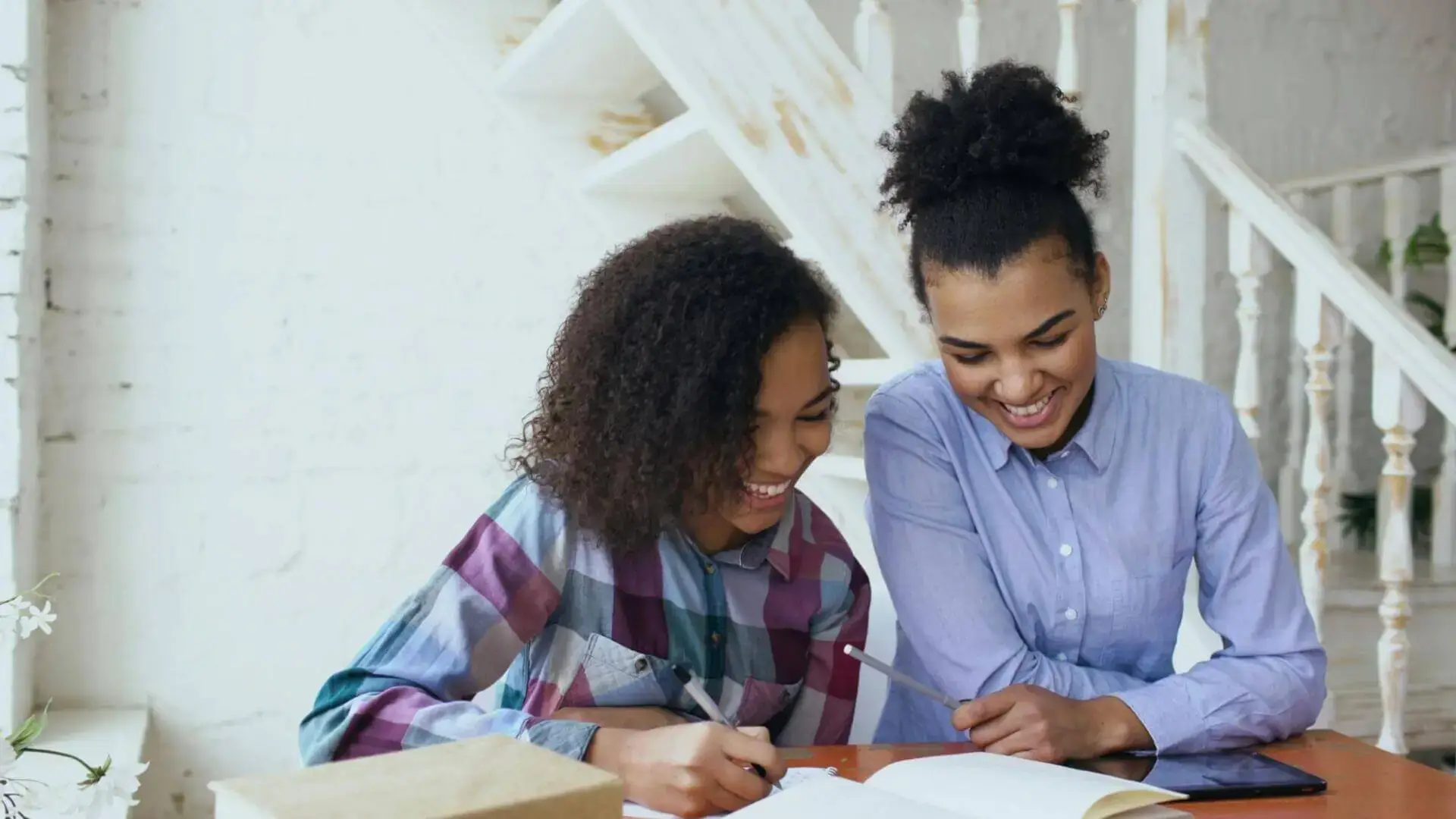 Two women sit at a table, smiling as they engage with an open notebook. The cozy, white background and natural light add a warm, cheerful ambiance.